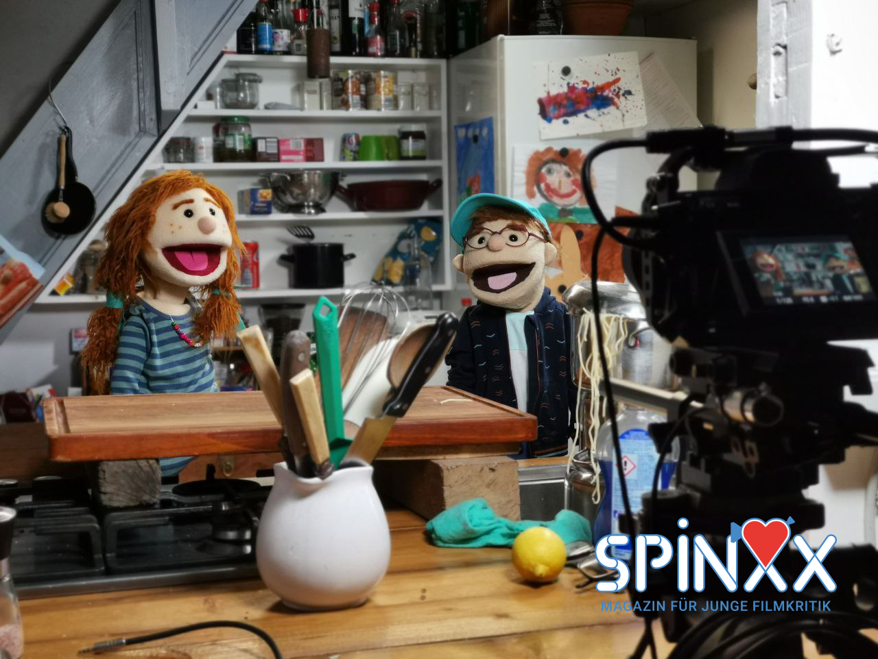 Puppen sitzen an einem Tisch und werden von einer Kamera gefilmt